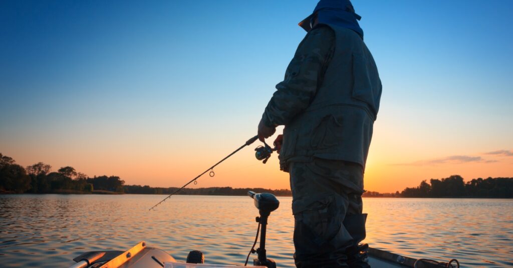 A fisherman standing on the stern of a fishing boat with a fishing rod at hand with the sun setting behind him.