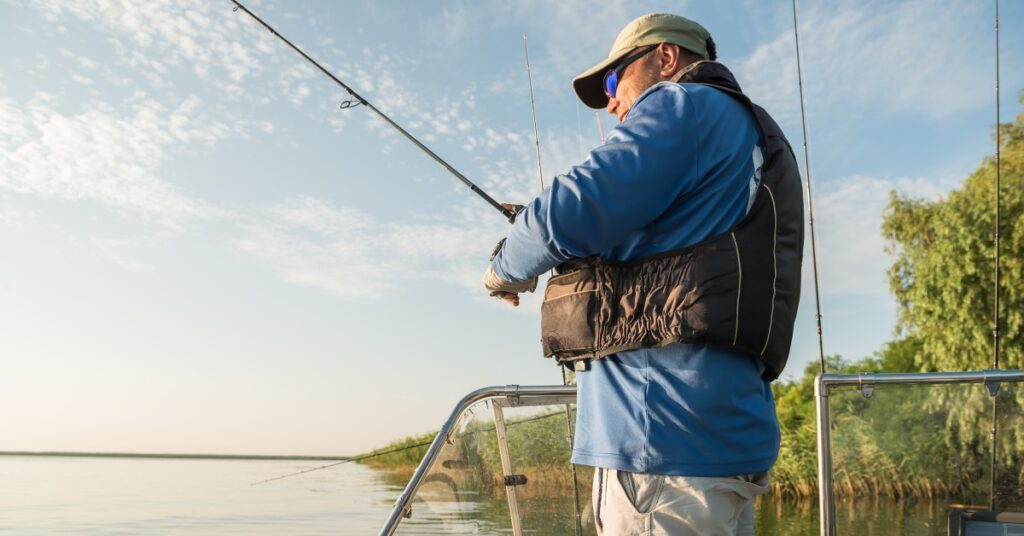 A professional angler wearing a life preserver fishing on a boat on a calm lake with another angler and road at the bow.