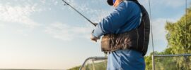 A professional angler wearing a life preserver fishing on a boat on a calm lake with another angler and road at the bow.