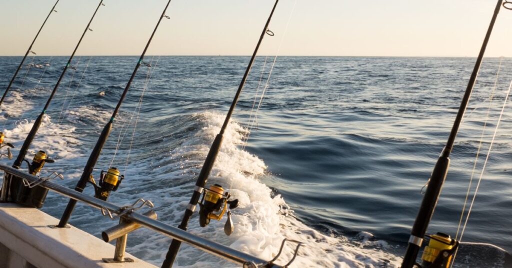 The port or starboard railing of a fishing charter boat with four fishing rods in holds looking out on the ocean.