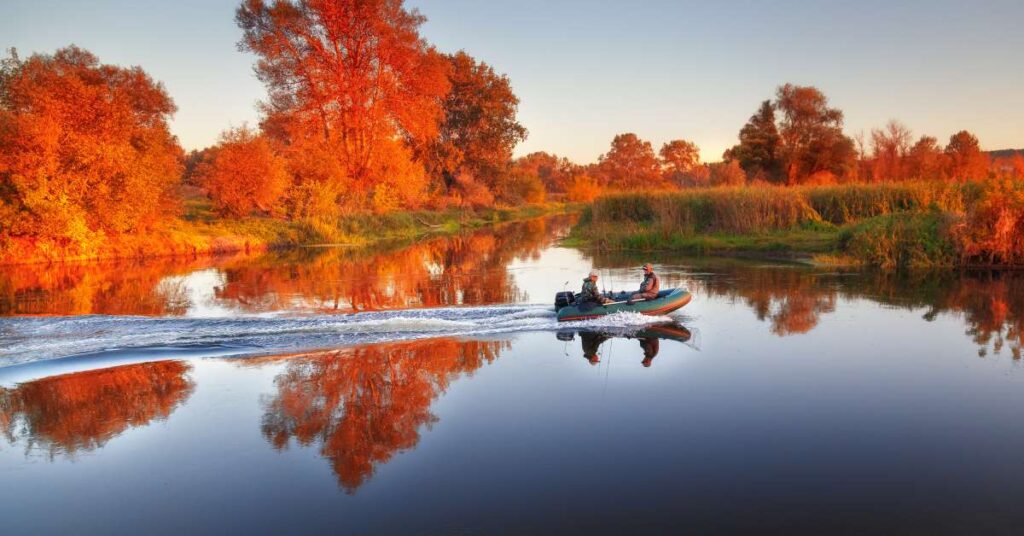 Two anglers sit on a rubber powerboat with fishing poles and gear on a river with fall tree foliage surrounding the river.