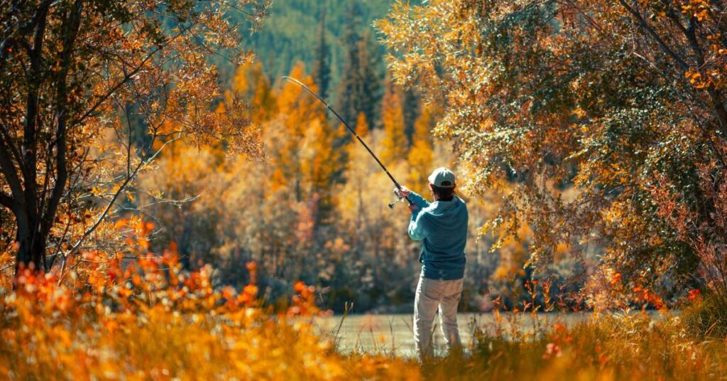 A man fishing holds a pole and reels in a catch while surrounded and framed by orange, red, and brown autumn tree leaves.