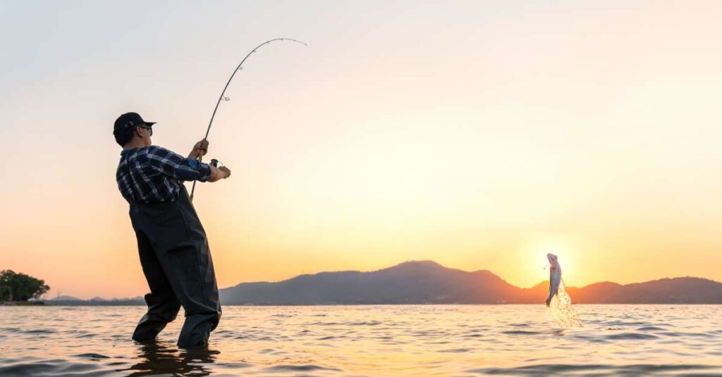 A male angler in waders and a flannel shirt pulls on his fishing rod, bending the rod and lifting a fish out of lake water.