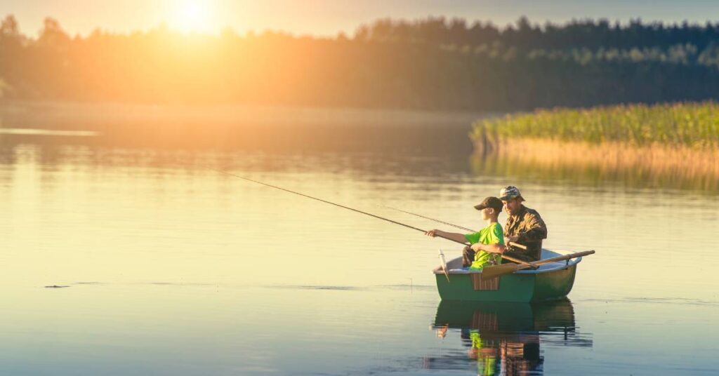 A man and a young boy with fishing poles fish in a canoe in the middle of a lake with the sun setting behind them.