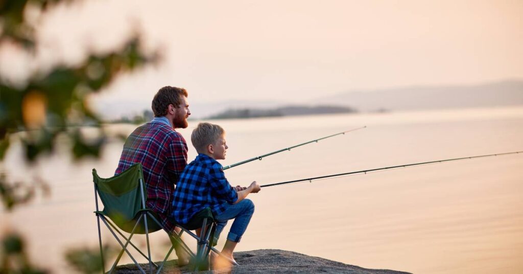 A man and a young boy sit on collapsible chairs on a rock overlooking a lake while holding fishing poles.