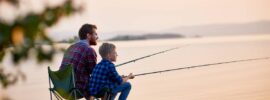 A man and a young boy sit on collapsible chairs on a rock overlooking a lake while holding fishing poles.