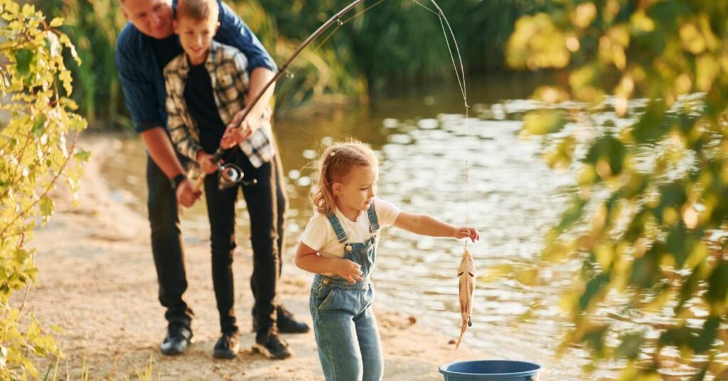 A man and a young boy use a fishing pole to lower a small fish into a bucket while a young girl holds the line.