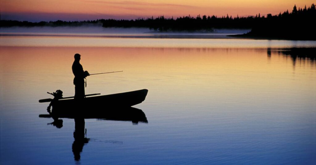 Silhouette of a man in a small fishing boat with a rod fishing in a lake at dawn on a lake with a forest in the background.