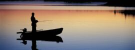 Silhouette of a man in a small fishing boat with a rod fishing in a lake at dawn on a lake with a forest in the background.