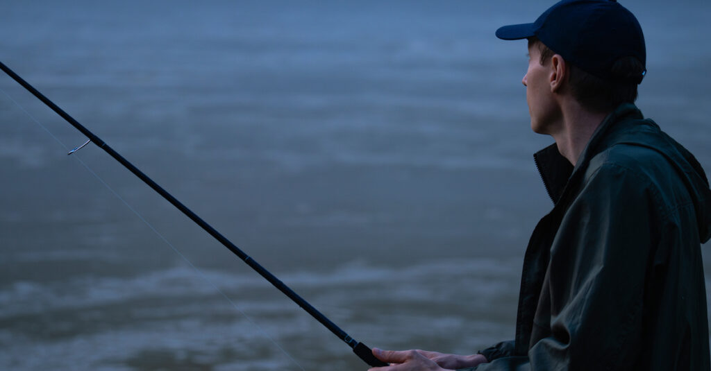 A fisherman wears a cap and raincoat and looks into a river while holding a fishing pole. It is nighttime.