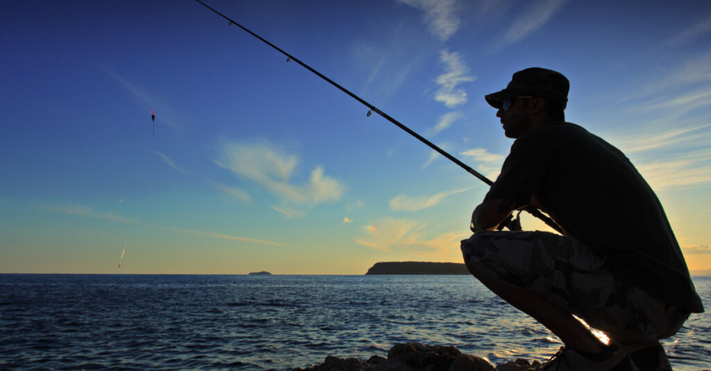 A man hunches down on a rock while holding a fishing pole next to a lake at sunset. He wears a cap and glasses.