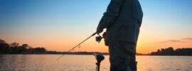 A man stands in a boat while fishing with his rod pointed down. In the background, the sun sets, and the sky gets darker.
