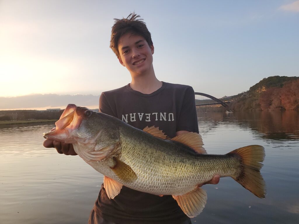 Young angler holding a largemouth bass caught on Lake Austin in December 2025 on a drop-shot finesse worm around submerged vegetation.
